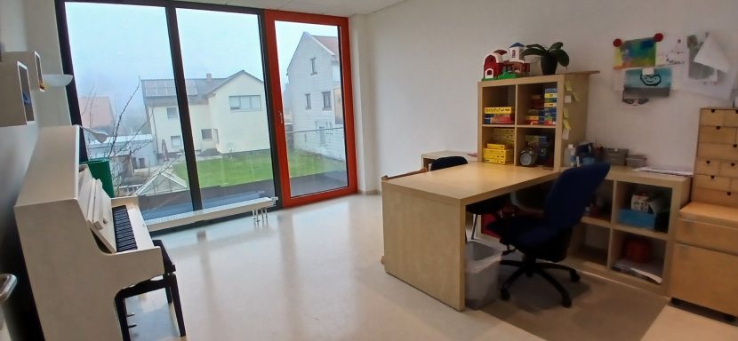 Raum 1 Raum 1: Heller Raum mit einem Schreibtisch, Regalen, einem Klavier und großen Fenstern.